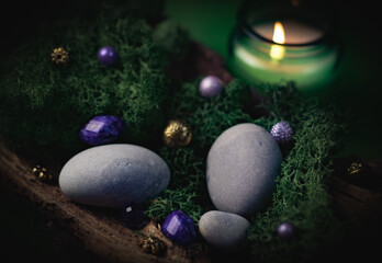 Lilac, golden stone lies in the bark of a tree with green moss, gray stones and a burning candle on...