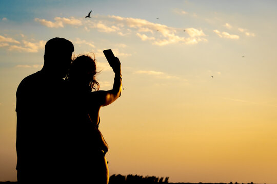 Silhouette of a couple embracing, taking a photo with their cell phone, of birds flying over the sea during a beautiful and romantic sunset. Concept of lovers, honeymoon, romance.