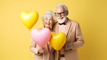Grandparents holding heart shaped balloons on pastel yellow background. A married couple of pensioners who are happy in love. Valentines Day composition.