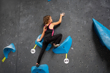 Little girl in sports clothes climbing wall of an entertainment centre. Child climbing wall at professional rock climb centre without use of hands. Child enjoys his hobby. Climb centre - obrazy, fototapety, plakaty