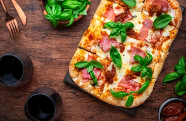 Foto auf Acrylglas Rectangular roman pizza with smoked ham, mozzarella cheese, sun dried tomatoes, tomato sauce and green basil leaves on rustic wooden table background, top view © 5ph