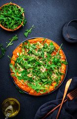 Hot homemade pizza with blue cheese, ham, pesto sauce and fresh arugula, black table background, top view
