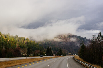 Asphalt road in the middle of high mountains, covered with fog and clouds on autumn day. American winter landscape of a mountainous area covered with fir forest. Fall season on the highway with cars