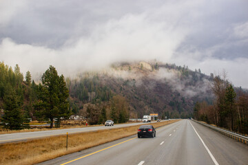 Asphalt road in the middle of high mountains, covered with fog and clouds on autumn day. American winter landscape of a mountainous area covered with fir forest. Fall season on the highway with cars