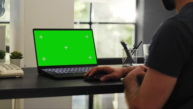 Male employee looks at greenscreen on laptop display, checking isolated chromakey layout in office coworking space. Businessman examining blank copyspace template at modern desk.
