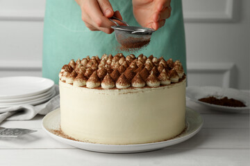Woman dusting delicious tiramisu cake with cocoa powder at white wooden table, closeup