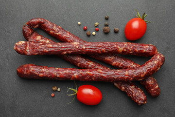 Many thin dry smoked sausages, peppercorns and tomatoes on black table, flat lay