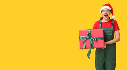 Beautiful female builder in Santa hat and with Christmas gift on yellow background with space for text