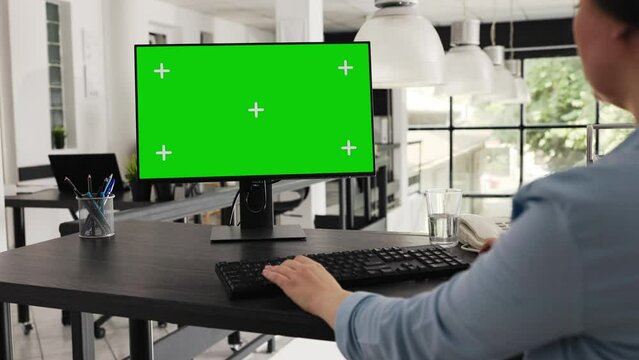 Employee sitting at desk with greenscreen on monitor, looking at isolated copyspace layout on computer desktop in coworking space. Businesswoman working on pc, blank mockup template.