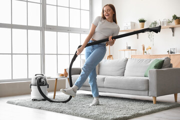 Young woman with vacuum cleaner singing at home - obrazy, fototapety, plakaty