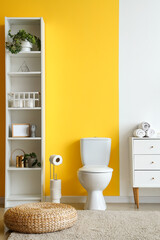Interior of modern restroom with ceramic toilet bowl, shelving unit and chest of drawers near yellow wall