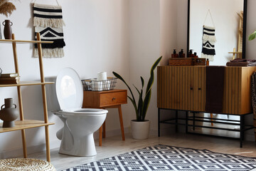 Interior of modern restroom with ceramic toilet bowl, wooden vanity cabinet and shelving unit