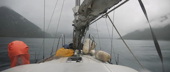Approaching the misty coast on sail yacht in low visibility on overcast rainy day, fog on the mountains, sail laid on boom, entering Russkaya bay, Kamchatka 
