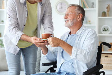 Senior man in wheelchair taking cup of tea from his son at home