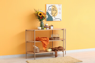 Wooden shelving unit with bouquet of autumn flowers, pumpkins and decor near orange wall