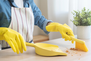 Household clean up, housekeeper asian young woman wearing protection rubber yellow gloves, using broom, dustpan sweeping remove spilled crumb food broken on dirty table at home, equipment for cleaning - obrazy, fototapety, plakaty
