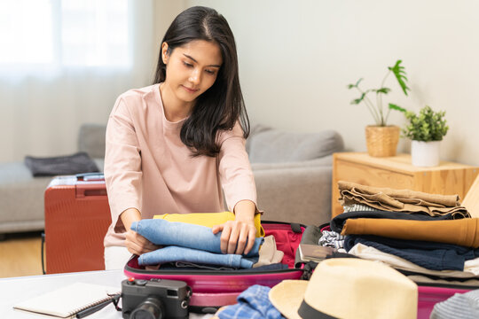 Travel concept, happy asian young woman hand preparing packing luggage bag for summer vacation trip on bed at home, pack her clothes and stuff, objects into bag suitcase for a new journey for leisure.