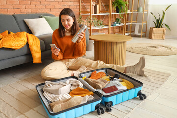 Young woman with mobile phone and thermos packing winter clothes in suitcase at home