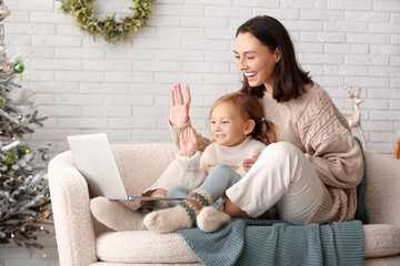 Happy young mother and her little daughter with laptop video chatting at home on Christmas eve