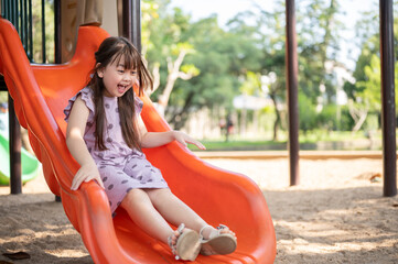 A playful and cheerful young Asian girl is playing a colorful slide in a playground in the park.