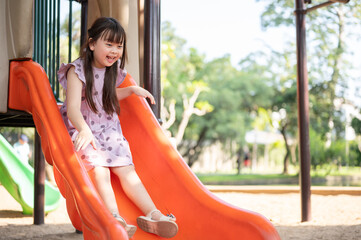 A playful and cheerful young Asian girl is playing a colorful slide in a playground in the park.