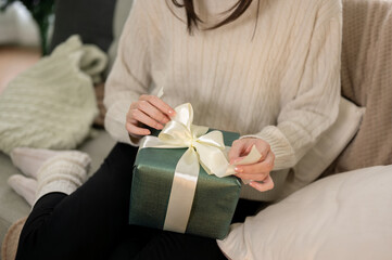 Close-up image of a woman is unwrapping a Christmas gift on a couch in her living room on Christmas - obrazy, fototapety, plakaty