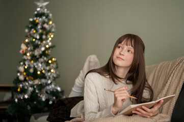 A beautiful woman is writing her diary or making a Christmas wish list while lying on a couch.