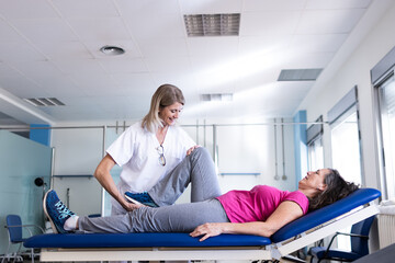 A middle aged blonde physiotherapist is rehabilitating her patient who is a brunette woman with leg...