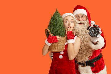Shocked people dressed as Santa Claus with Cypress tree and alarm clock on orange background