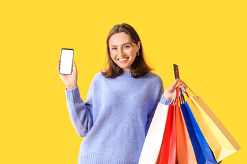 Young woman with credit card, mobile phone and shopping bags on yellow background - obrazy, fototapety, plakaty