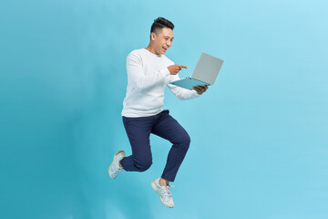 Cheerful asian young man holding laptop smiling jumping high running isolated on blue background.