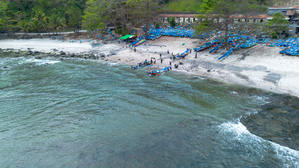 Drone view of Menganti Beach with white sand, calm waves, cliffs and green trees and the activities of traditional fishermen and traditional fishing boats