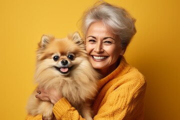 Portrait of smiling Caucasian senior woman in yellow sweater posing with cute Pomeranian spitz puppy. Happy lady hugs her beloved pet. Love between human and dog. Yellow studio background, copy space.