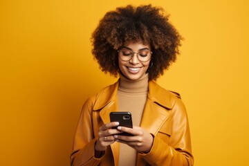 Studio portrait of beautiful African American girl with smartphone in yellow clothes against yellow background. Smiling lady with Afro hairdo texting message, enjoying online communication, using app.