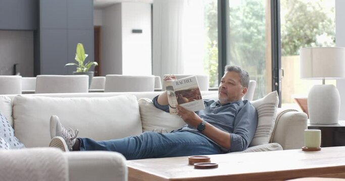 Focused senior biracial man relaxing on couch reading book in living room, slow motion