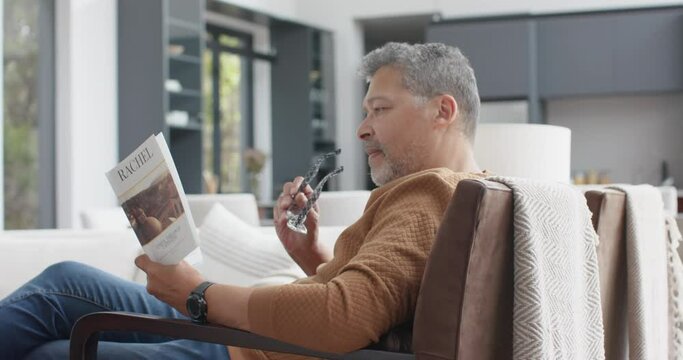 Focused senior biracial man sitting in armchair reading book in living room, slow motion
