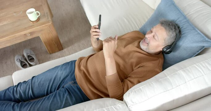 Happy senior biracial man on couch in headphones listening to music on smartphone, slow motion