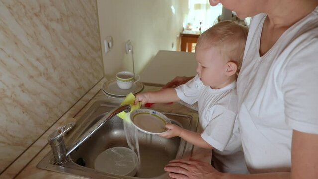 Little daughter washes dishes with her mother in the kitchen.Hands of a child who takes dishwashing detergent and a dishwashing sponge. Mom teaches child to wash dishes in the kitchen. 