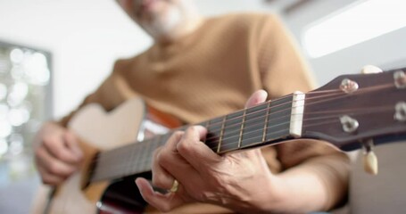 Midsection of senior biracial man sitting playing guitar at home, selective focus, slow motion