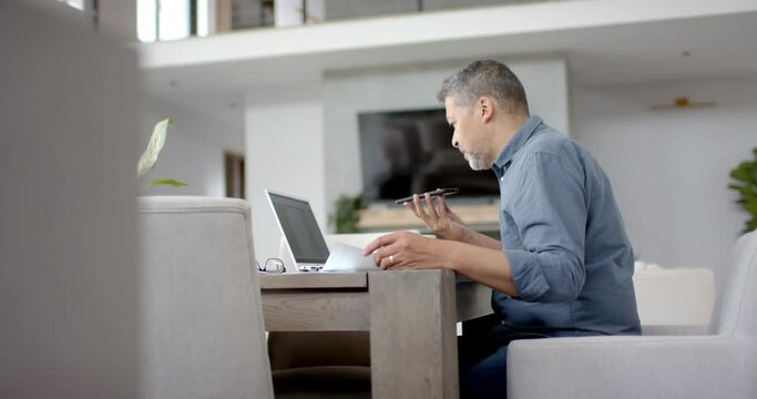 Senior biracial man using smartphone and laptop at desk, working from home, copy space, slow motion