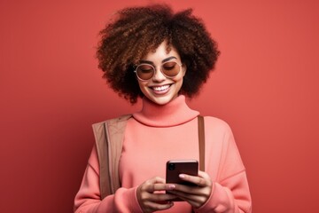 Fototapeta na wymiar Studio portrait of beautiful African American woman with smartphone in pink clothes against pink background. Positive girl with Afro haircut texting message, enjoying online communication, using app.