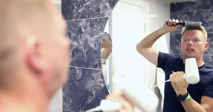Young man standing near mirror and combing his hair with hairdryer in bathroom 4k movie. Home hair care for men concept