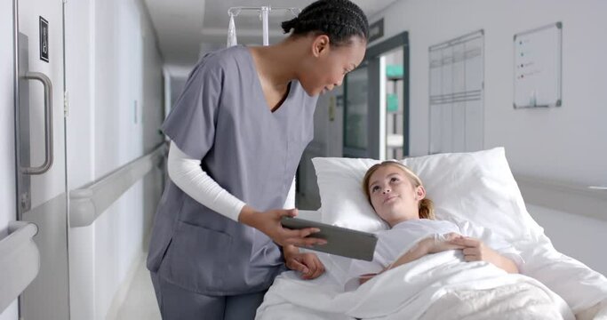 Diverse female doctor and girl patient lying on bed using tablet in hospital, slow motion