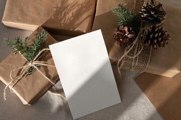 Christmas holiday presents and postcard mockup on table. Brown crafted gift boxes decorated with pine cones, blank paper card template on neutral beige linen table background with sunlight shadow