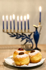 Beautiful Menorah (Chanukkiah) with 8 lit burning candles for Jewish Hanukkah holiday on table at home. Celebrating Chanukah festival of lights.  Sufganiyot donuts sweet cultural food on a plate.