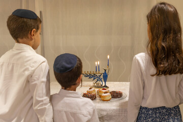 Three children looking at the Menorah with 8 lit burning candles for Jewish Hanukkah holiday on table at home. Celebrating Chanukah festival of lights. Dreidel and Sufganiyot donuts on a plate. 