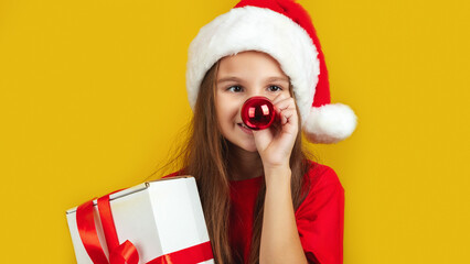 Holiday Fun. Cheerful Little Girl Wearing Santa hat Holding Christmas Present, Playful Kid Keeping Xmas Tree Ball Near Nose And Looking At Camera, Yellow Background