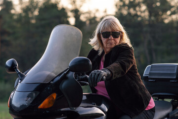 Stylish blonde motorcycle tourist, super mom in black gloves, black glasses, sitting on her bike.
