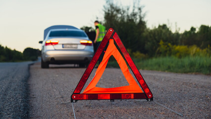 Broken silver luxury car emergency accident. Man driver installing red triangle stop sign on road....