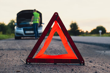 Unrecognizable sad driver in reflective vest. Male driver standing near broken car with open up hood. Red triangle to warn other road users of car breakdown or engine failure stop at countryside